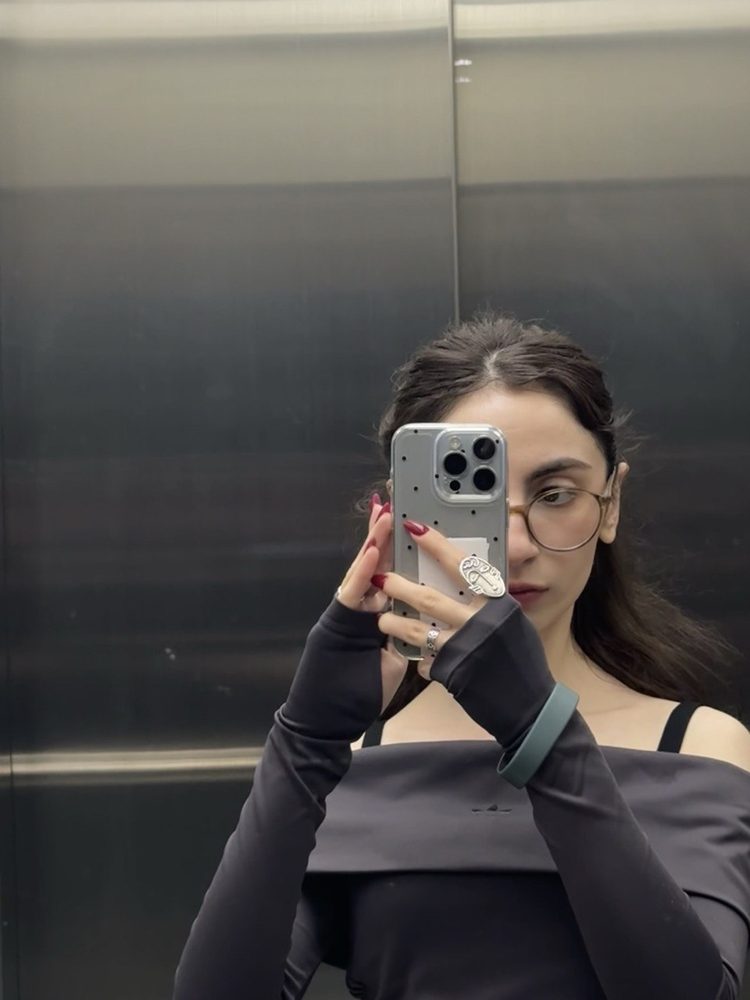 Yevgenya taking an elevator mirror selfie in glasses and a black off-shoulder top, red nails on the phone.